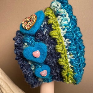 May include: A blue, green, and white crocheted hat with a heart-shaped button and three pink heart-shaped buttons.