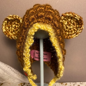 May include: A brown crocheted monkey hat with yellow trim. The hat has a hood and ear flaps.