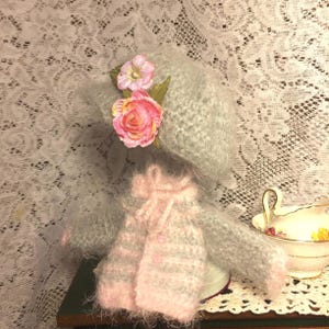 Può includere: Una bambola di lana grigia e rosa con un cappello grigio e fiori rosa. La bambola indossa un maglione a righe rosa e grigio.