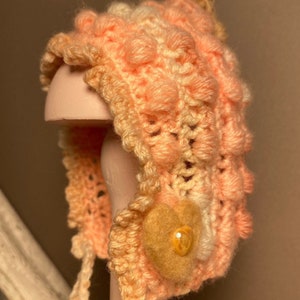 May include: A crocheted baby bonnet in shades of peach and cream with a textured, puffy design. The bonnet has a small, felt flower embellishment with a yellow button centre.