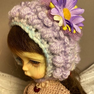 May include: A doll wearing a purple and blue crocheted hat with a purple flower on top. The doll is wearing a brown knitted sweater.