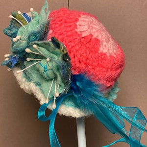 Puede incluir: Un sombrero de crochet con un diseño rosa y blanco, adornado con una pluma azul y una flor verde y azul con detalles blancos. El sombrero está atado con una cinta azul.