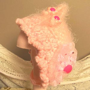 May include: A pink crocheted hat with a fuzzy texture and pink flower embellishments. The hat has a scalloped edge and a sheer pink fabric overlay.