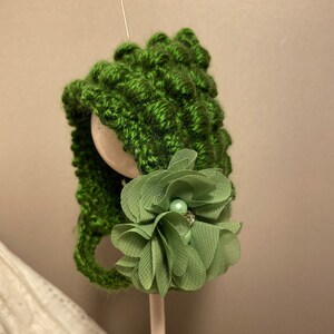 Peut inclure: Un bonnet de bébé tricoté à la main, vert, avec un motif texturé. Le bonnet est orné d'une fleur en tissu vert sauge et d'un accent de perle. Le bonnet est présenté sur une forme de tête en bois.