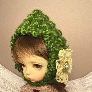 May include: A doll wearing a green crocheted hat with a white crocheted flower accent. The flower has a pearl-like button in the center.