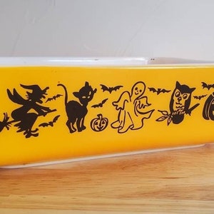 May include: A yellow ceramic baking dish with a Halloween theme. The dish features black silhouettes of a witch on a broomstick, a cat, a ghost, an owl, and a jack-o'-lantern.