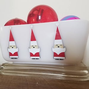 May include: White ceramic butter dish with a clear glass lid. Three Santa Claus figures are painted on the front of the dish. The dish is filled with red and pink Christmas ornaments.