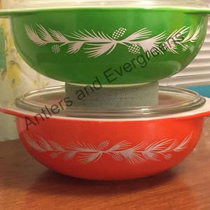 May include: Two vintage Pyrex mixing bowls with lids. The top bowl is green with a white pine bough design. The bottom bowl is orange with a white pine bough design. Both bowls have a white rim.
