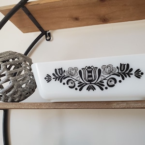 May include: A white ceramic baking dish with a black floral design. The dish is sitting on a wooden shelf.