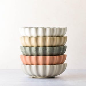 May include: A stack of five round bowls with a scalloped, fluted edge design. The bowls are colored in a gradient of white, light beige, muted sage green, soft terracotta orange, and a light grey, each featuring a smooth, matte finish.