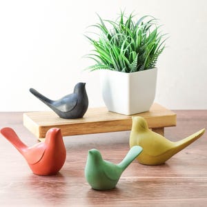 May include: A collection of colorful bird figurines in red, green, yellow, and black. The birds are arranged on a wooden surface with a small wooden shelf holding a white square pot with a green plant.