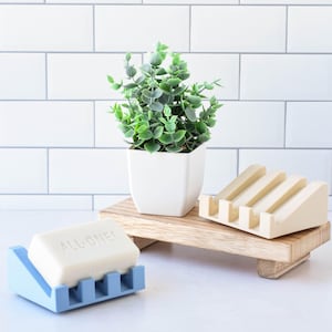 May include: A light blue soap dish with a bar of soap that reads "ALL-ONE!". A beige soap dish and a small potted plant sit on a wooden tray. The background is a white tiled wall.