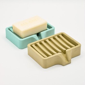 K&ouml;nnte beinhalten: Zwei Seifenschalen, eine hellt&uuml;rkis und eine beige, werden gezeigt. Die t&uuml;rkisfarbene Schale enth&auml;lt ein St&uuml;ck Seife. Die beige Schale hat ein Latten-Design. Beide sind aus einem festen Material.