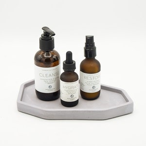 May include: Three glass bottles with black pump tops and labels on a light gray concrete tray. The labels read "Cleanse", "Hydrate", and "Restore".