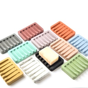 May include: Assortment of 10 colorful soap dishes made of concrete. Each dish has a rectangular shape with a raised, ridged design. The dishes are in various colors, including gray, green, blue, pink, orange, and white.