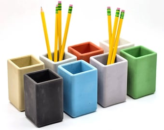 May include: Set of eight small, square concrete pencil holders in various colors, including beige, gray, blue, light gray, orange, and green. The holders are arranged in a row on a white surface.