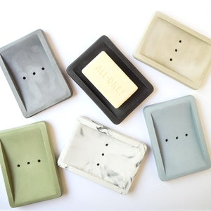 May include: Six rectangular soap dishes made of concrete in various colors, including gray, green, white, and blue. One dish is black and features a bar of soap with the text "ALL-ONE!" printed on it.