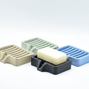 K&ouml;nnte beinhalten: Vier Seifenschalen aus Beton in Beige, Gr&uuml;n, Schwarz und Blau. Eine Schale enth&auml;lt ein St&uuml;ck Seife. Jede Schale hat eine rechteckige Form mit einem geschlitzten Design und einem kleinen Ausguss.