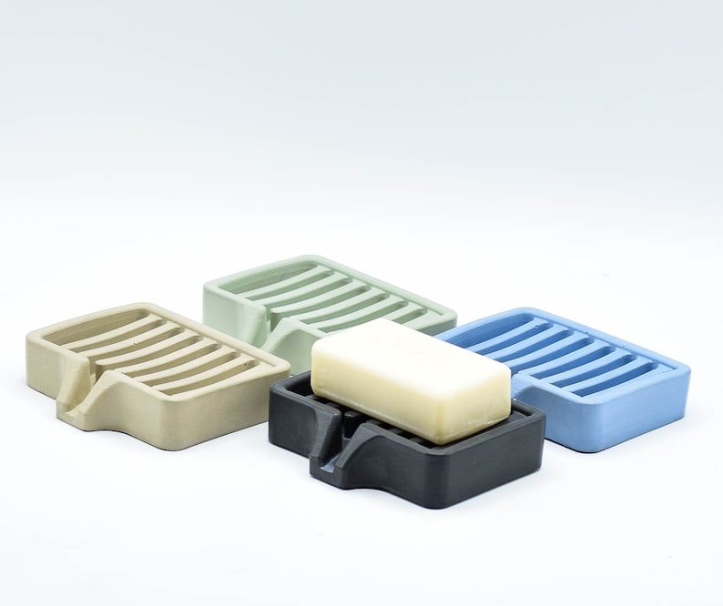 K&ouml;nnte beinhalten: Vier Seifenschalen aus Beton in Beige, Gr&uuml;n, Schwarz und Blau. Eine Schale enth&auml;lt ein St&uuml;ck Seife. Jede Schale hat eine rechteckige Form mit einem geschlitzten Design und einem kleinen Ausguss.