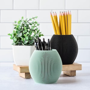 May include: A decorative desk arrangement featuring a small potted plant, a light green ribbed pen holder, and a black ribbed pencil holder. The pencil holder is filled with yellow pencils. The items are on wooden platforms.