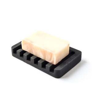 May include: A black silicone soap dish with ridges to help drain water. A bar of white soap with swirls of pink is resting on the dish.
