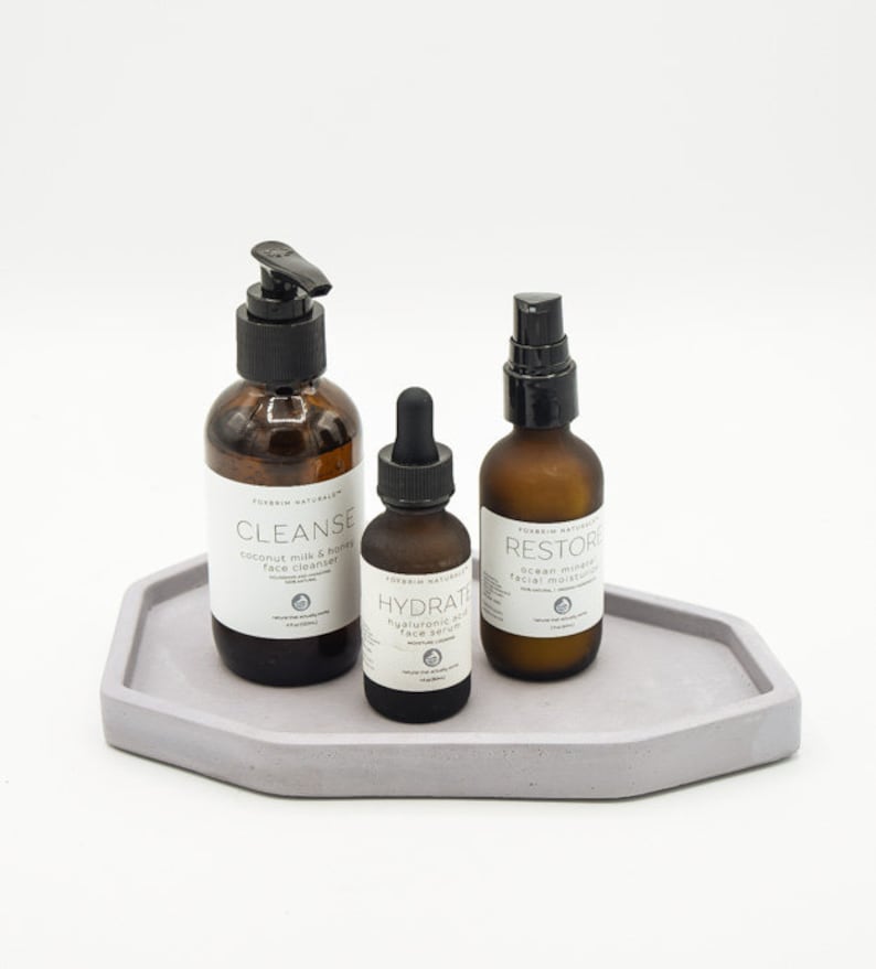 May include: Three glass bottles with black pump tops and labels on a light gray concrete tray. The labels read "Cleanse", "Hydrate", and "Restore".