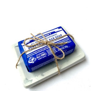 May include: A blue bar of Dr. Bronner's Pure Castile Peppermint soap with organic oils, wrapped in twine and sitting on a gray concrete soap dish. The soap is labeled "All-One!" and "Certified Fair Trade".