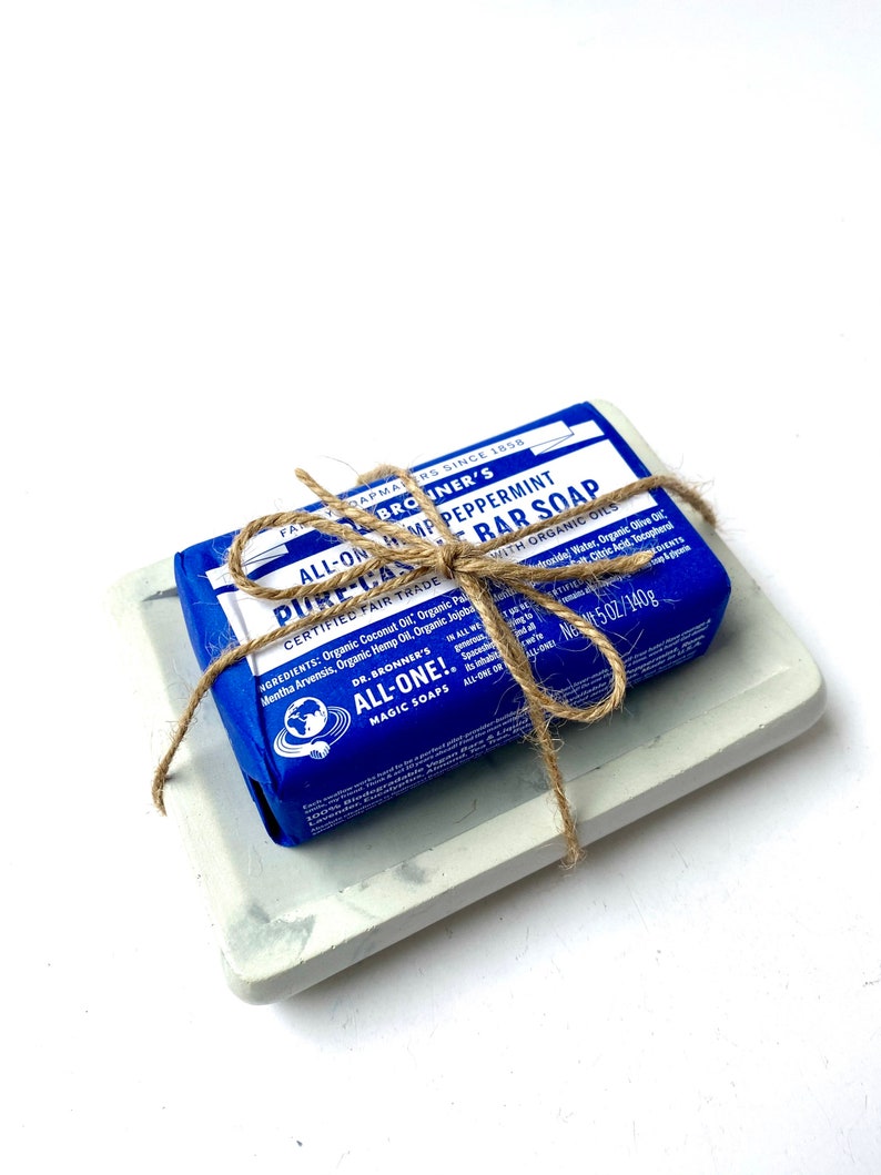 May include: A blue bar of Dr. Bronner's Pure Castile Peppermint soap with organic oils, wrapped in twine and sitting on a gray concrete soap dish. The soap is labeled "All-One!" and "Certified Fair Trade".