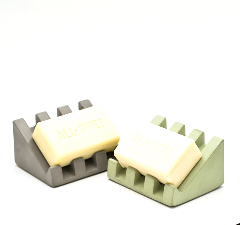 May include: Two soap dishes made of concrete, one gray and one green, each with a bar of soap. The soap on the gray dish says "ALL-ONE!" and the soap on the green dish says "ALL-ONE!"