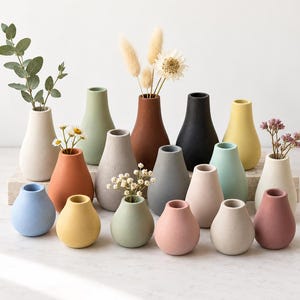 May include: A collection of small, matte-finish ceramic vases in various solid colors including blue, yellow, green, pink, terracotta, gray, black, and white. The vases have a rounded, teardrop-shaped body with a narrow neck and open top.
