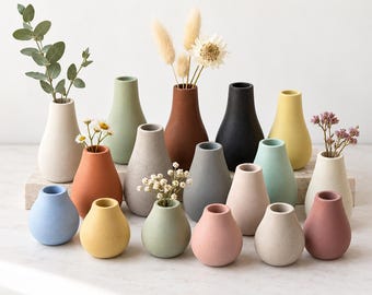 May include: A collection of small, matte-finish ceramic vases in various solid colors including blue, yellow, green, pink, terracotta, gray, black, and white. The vases have a rounded, teardrop-shaped body with a narrow neck and open top.