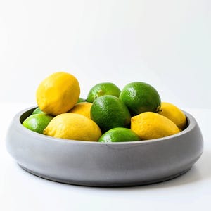 May include: A shallow, matte gray circular bowl filled with a collection of bright yellow lemons and vibrant green limes. The fruit is piled together in the center of the container, creating a contrast between the citrus colors and the neutral bowl.