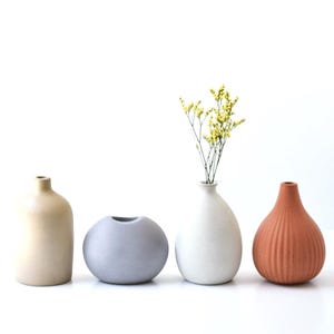 May include: Four ceramic vases in a row, each with a unique shape and color. The first is beige, the second is gray, the third is white with yellow flowers, and the fourth is terracotta. A minimalist home decor item.
