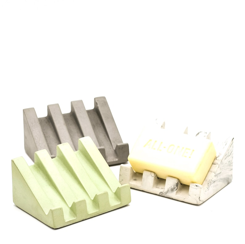 May include: Three soap dishes made of concrete in gray, green, and white with gray marbling. The white dish has a bar of soap on it that says "ALL-ONE!" in yellow lettering.
