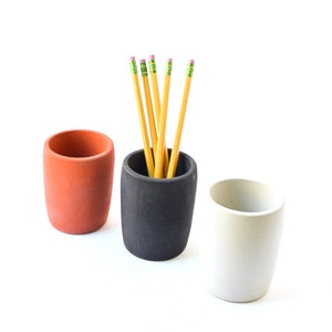 May include: Three cylindrical pencil holders in orange, black, and white. The black holder contains several yellow pencils with pink erasers. The holders are made of a matte material and are arranged on a white background.
