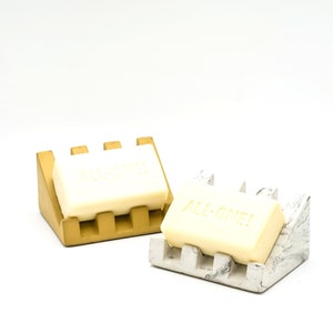 May include: Two soap dishes, one gold and one white with gray marbling, each holding a bar of soap. The soap on the gold dish has the word "ALL-ONE!" embossed on it.