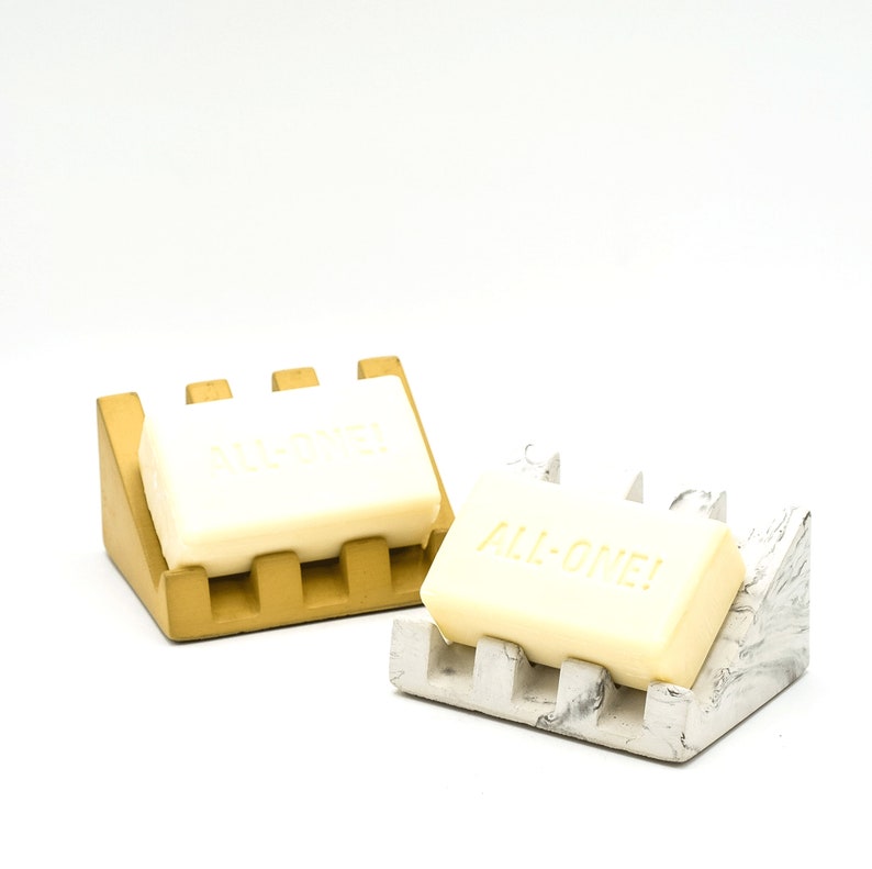 May include: Two soap dishes, one gold and one white with gray marbling, each holding a bar of soap. The soap on the gold dish has the word "ALL-ONE!" embossed on it.