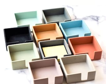May include: Ten square note holder stands in various colors, including black, green, gray, blue, orange, and beige. The stands are made of a smooth, textured material and have a slot for holding sticky notes.