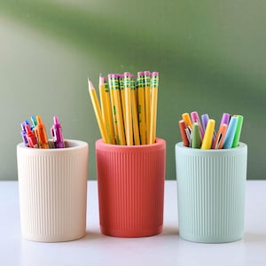 May include: Three cylindrical pencil holders in cream, coral, and mint green, each with vertical ribbed detailing. The holders contain pencils and pens in various colors, perfect for a desk or office setting.