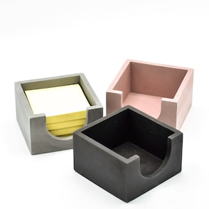 May include: Three concrete note holders, one gray, one pink, and one black. The gray holder has a stack of yellow sticky notes inside.