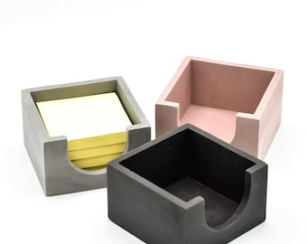 May include: Three concrete note holders, one gray, one pink, and one black. The gray holder has a stack of yellow sticky notes inside.