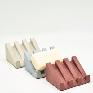 May include: Three soap dishes made of concrete in different colors: beige, light blue, and burgundy. The beige and light blue dishes are empty, while the burgundy dish has a bar of soap on it. The soap is white and has the word "ALL-OVER" printed on it.