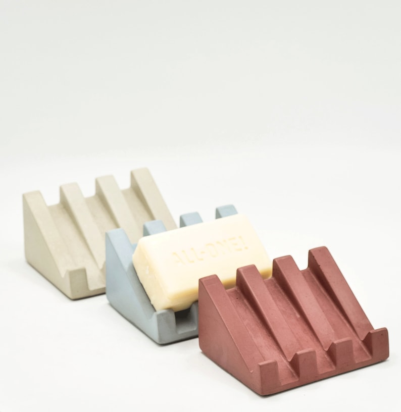 May include: Three soap dishes made of concrete in different colors: beige, light blue, and burgundy. The beige and light blue dishes are empty, while the burgundy dish has a bar of soap on it. The soap is white and has the word "ALL-OVER" printed on it.