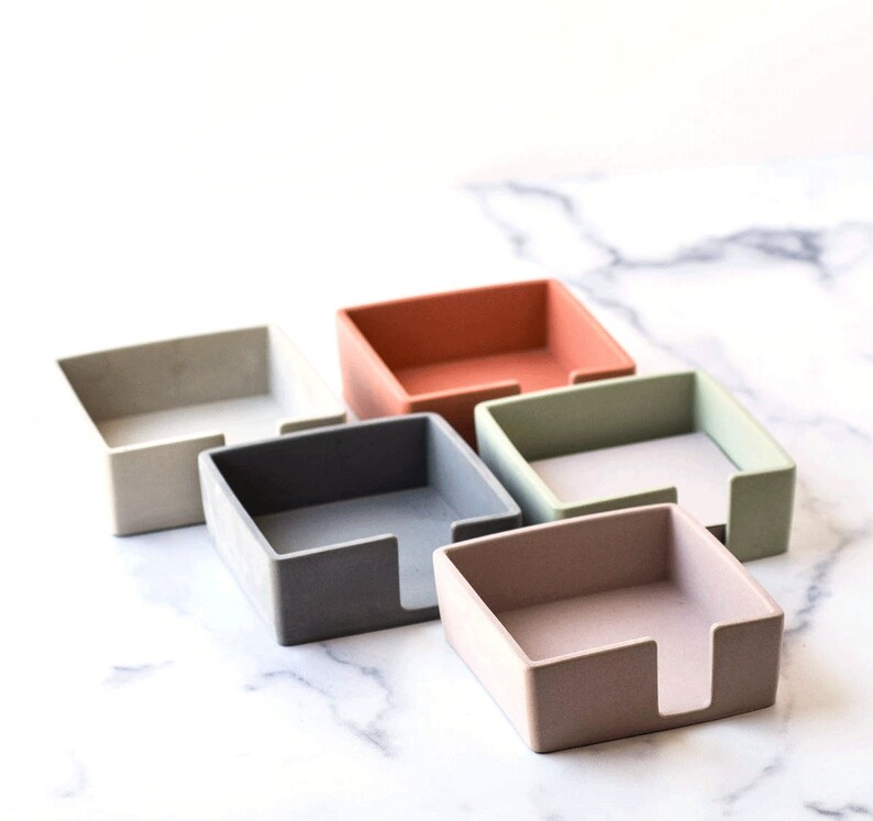 K&ouml;nnte beinhalten: F&uuml;nf quadratische Notizhalter in verschiedenen Farben: Grau, Hellgrau, Hellgr&uuml;n, Hellrosa und Orange. Die Halter bestehen aus einem strukturierten Material und haben einen Schlitz zum Halten von Notizen.