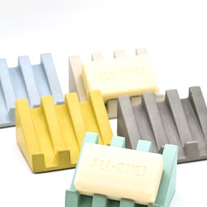 May include: Five soap dishes in different colors: light blue, beige, yellow, gray, and light green. Each dish has a raised design and is made of concrete. A bar of soap with the text "ALL-ONE!" is on the light green dish.