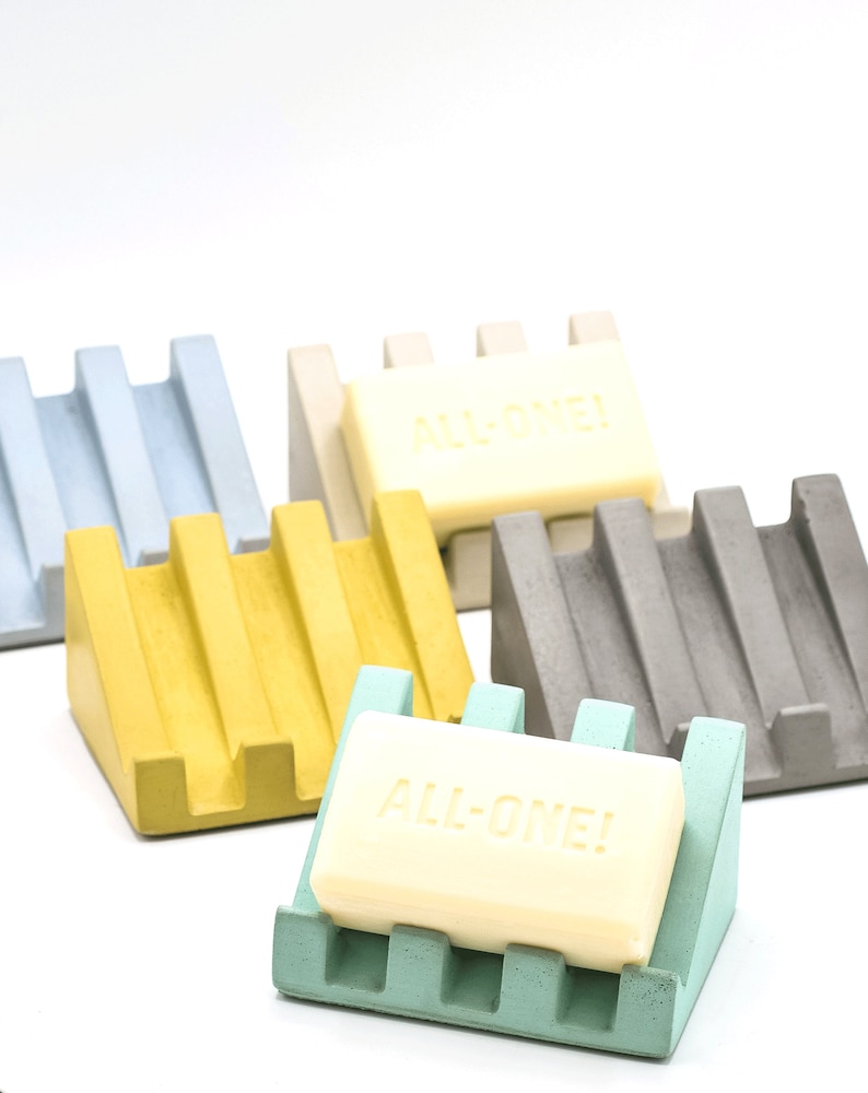 May include: Five soap dishes in different colors: light blue, beige, yellow, gray, and light green. Each dish has a raised design and is made of concrete. A bar of soap with the text "ALL-ONE!" is on the light green dish.