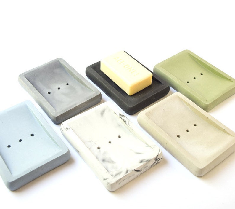 May include: Six different soap dishes made of concrete in various colors, including gray, blue, white, black, green, and beige. The dishes have a curved shape and three drainage holes. A bar of soap is sitting on one of the dishes.
