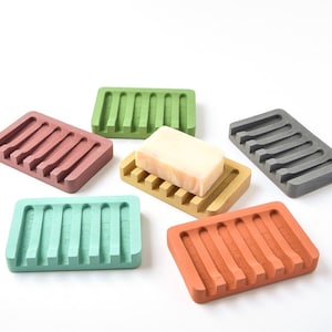 May include: Six colorful soap dishes made of a textured material with ridges. The dishes are in shades of green, brown, gray, orange, and pink. A bar of soap is sitting on one of the dishes.