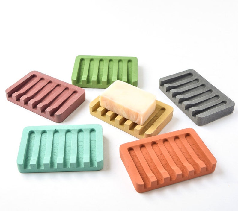 May include: Six colorful soap dishes made of a textured material with ridges. The dishes are in shades of green, brown, gray, orange, and pink. A bar of soap is sitting on one of the dishes.