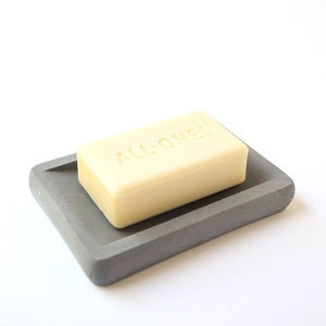 May include: A gray concrete soap dish with a rectangular shape and rounded edges. A bar of soap with the words "ALL-ONE!" embossed on it is sitting on the dish.